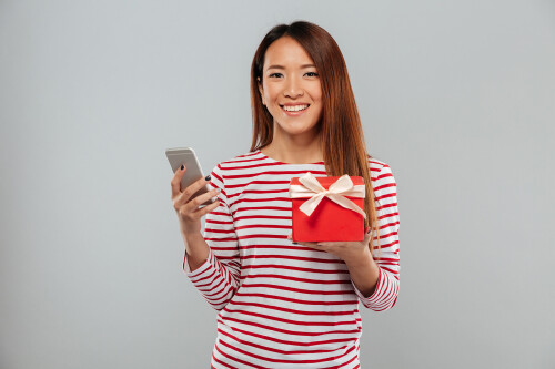 Image of cheerful young asian woman chatting by phone standing isolated over grey wall. Looking camera holding gift.